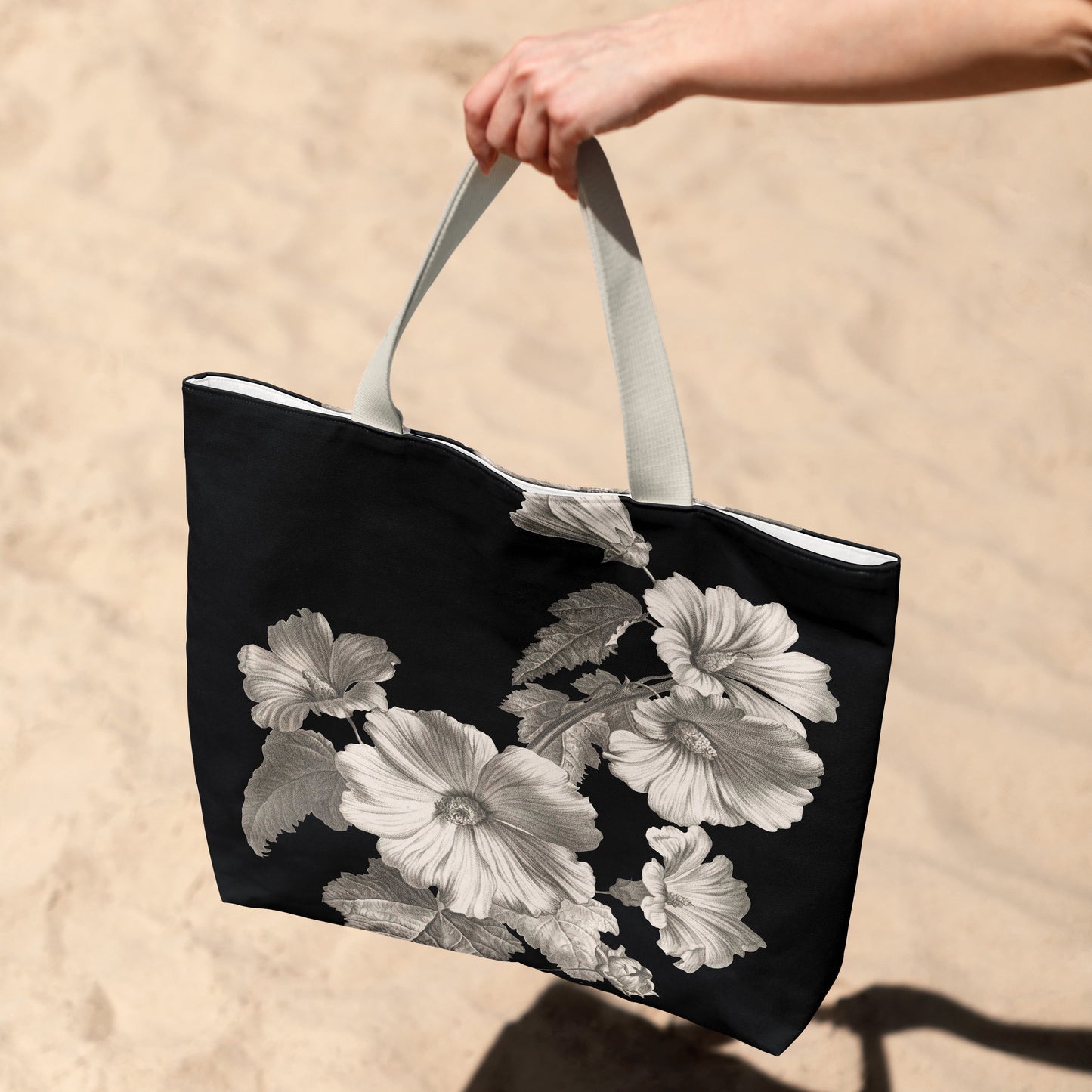 Black tote bag with white floral design held by a hand on a sandy background